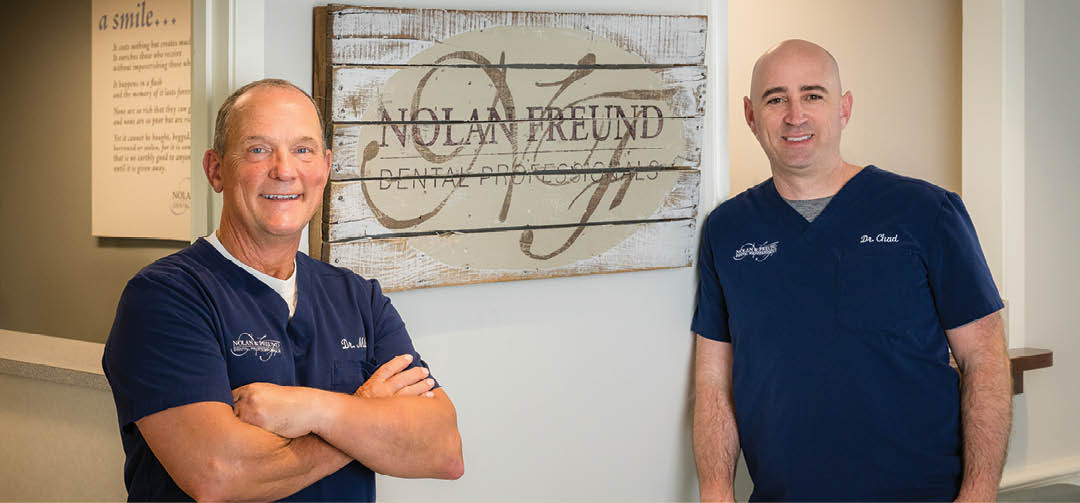 Two men in blue shirts standing in front of a sign. Description generated by AI
