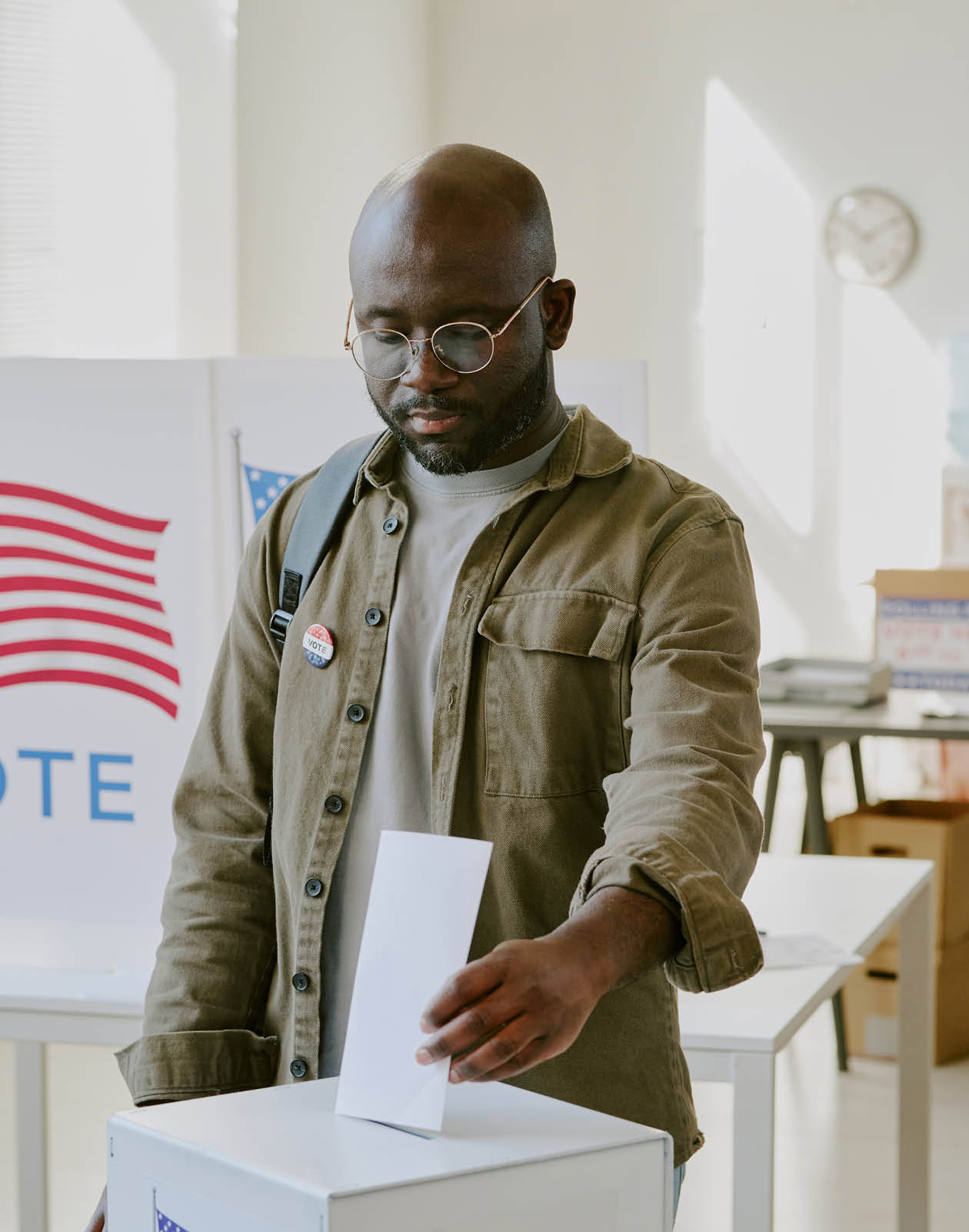 Vertical medium shot of young African American male citizen putting ballot paper into ballot box at polling place on election day