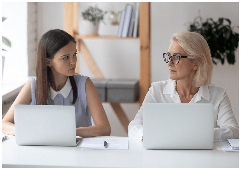 Different ages women colleagues 50s and young female workmates sit at office desk looking with enmity at each other. Negative attitude, conflict at workplace, misunderstanding generational gap concept