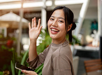 A portrait of a beautiful, positive Asian woman in casual clothes stands outdoors with a smartphone, waves and smiles at the camera. people, lifestyles, and wireless technology