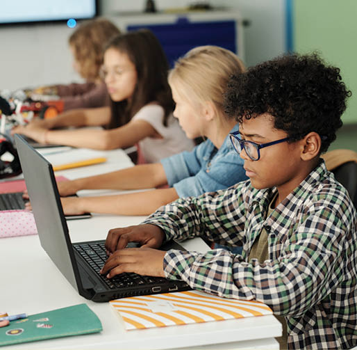 Side view of youthful African American schoolboy and his classmates working in front of laptops while sitting in row by desk