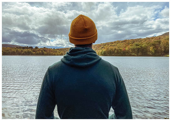 In crisp autumn weather, a sweater and beanie keep a millennial young man warm as he peacefully stares at the lake. The Catskill mountains are one of the most beautiful places in the United States for camping, hiking, and seeing fall foliage.