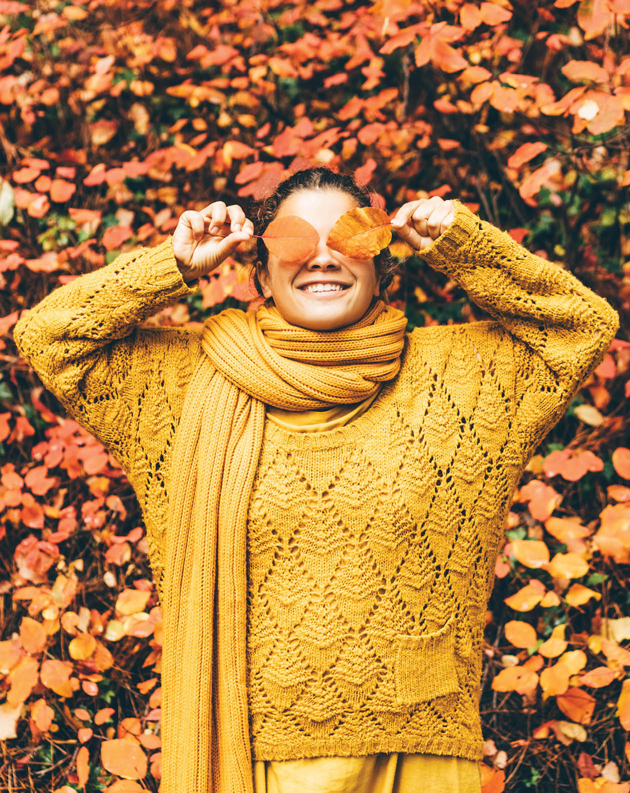 Playful young woman in bright orange clothes with long scarf hides eyes behind dry orange leaves near large red bush in picturesque autumn forest