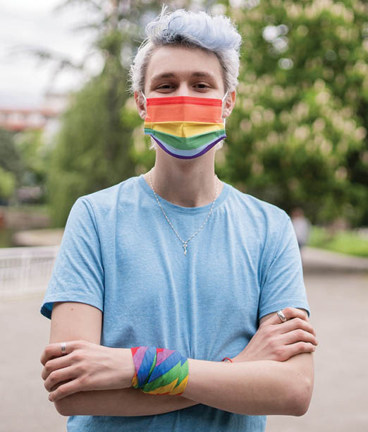 A man with pale blue dyed hair is standing ouside with his arms crossed, wearing a blue t-shirt and a rainbow striped protective face mask on a cloudy day in a park