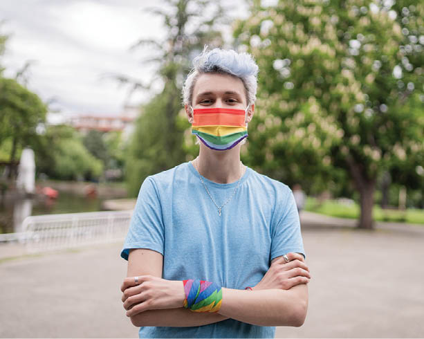 A man with pale blue dyed hair is standing ouside with his arms crossed, wearing a blue t-shirt and a rainbow striped protective face mask on a cloudy day in a park