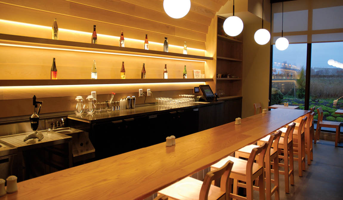 A bar with a wooden counter and a row of chairs. Description generated by AI