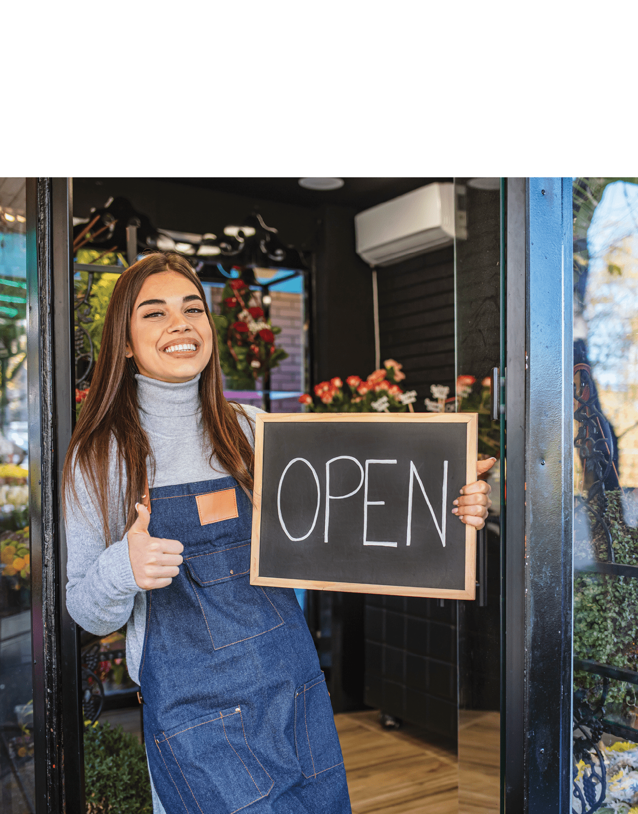 Woman inviting to her flower shop  Beautiful smiling young florist in apron holding open sign  Smiling woman holding open sign, written on blackboard with chalk, outside of flower shop 