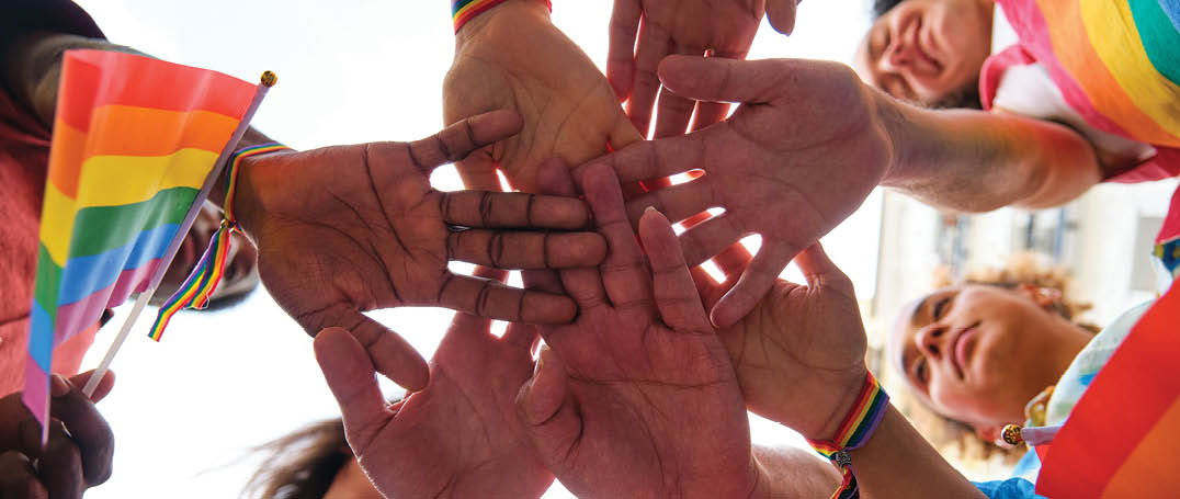 A group of multiracial people with rainbow LGBTQ bracelets stacking hands. Scene is one of unity and support for the LGBTQ community.