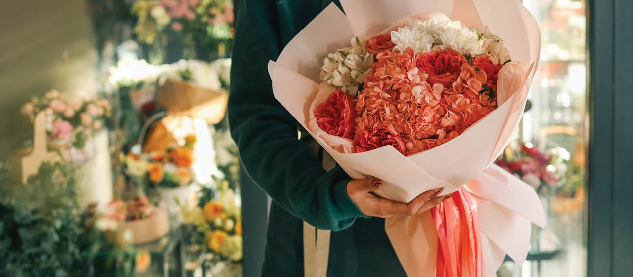 Elegant bouquet with hydrangeas, roses, and delicate flowers wrapped in soft pastel paper with bright ribbons, held by a florist in a cozy flower shop, highlighting vibrant blooms.