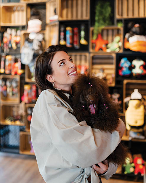 Young woman smiling as she hugs her affectionate brown poodle while shopping for treats and accessories in a bright, modern pet supply boutique, enjoying their close bond