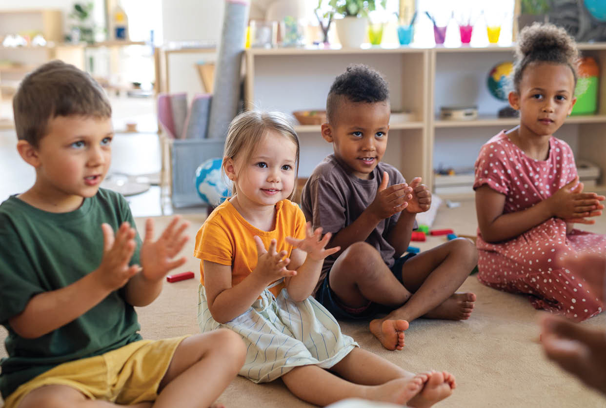 A group of small nursery school children sitting on floor indoors in classroom, clapping.