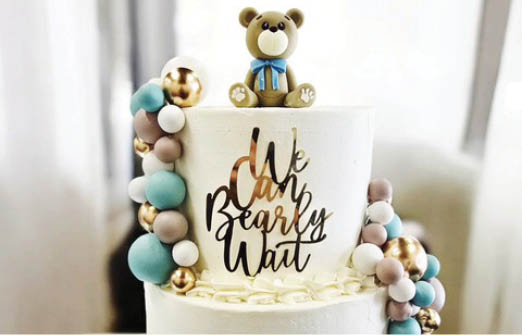 A white cake with gold and blue decorations and a gold teddy bear on top. Description generated by AI