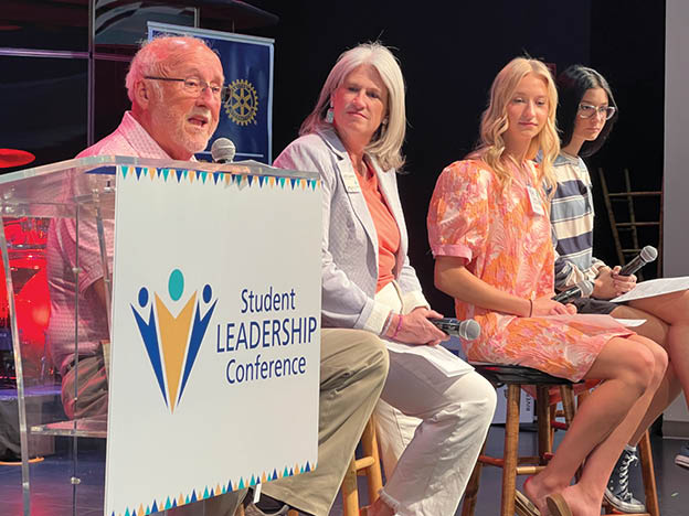 A man is speaking at a podium with three women sitting in chairs behind him. Description generated by AI