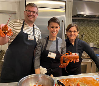 Three people are standing in a kitchen, smiling and holding up cooked lobsters. Description generated by AI