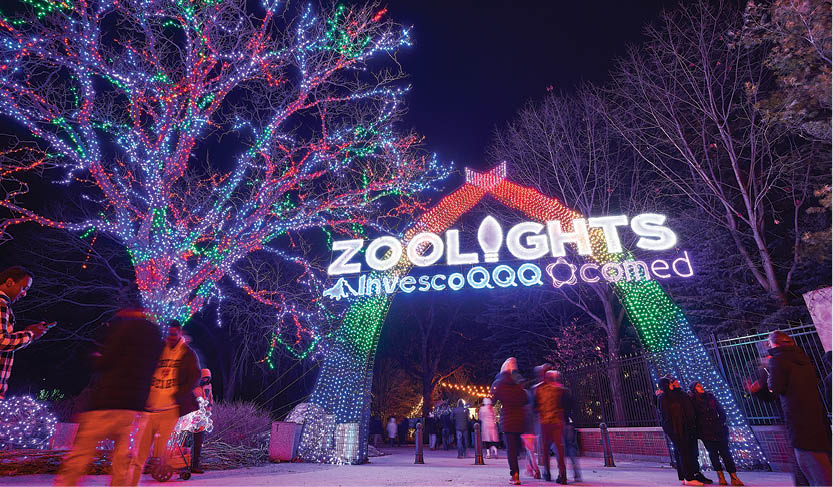 A group of people standing in front of a sign that says ZooLights. Description generated by AI