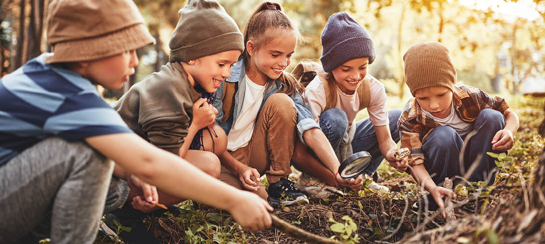 Happy children boys and girls in casual clothes with backpacks making bonfire with magnifying glass together in green forest during school camping activity on sunny day, smiling kids exploring nature