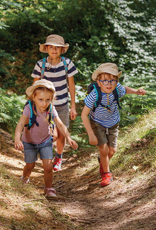 Three young adventurers, equipped with hats and backpacks, trek along a forested path, greenery all around