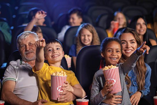 Front view of multiracial kids holding popcorn and sitting on knees of parents. Little african girl with caucasian woman and caucasian boy with senior grandfather. Family, entertainment concept.