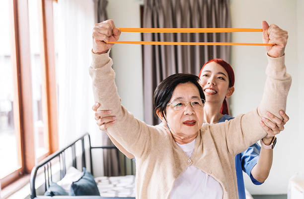 Nurse caregiver wearing scrubs exercises with a senior Asian woman by using resistance band exercise for the senior patient in physiotherapy treatment. Home health care and nursing home concept.