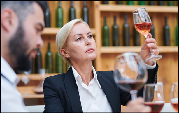 Confident blonde female sommelier or cavist looking at color of wine in wineglass at work in cellar