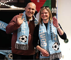 A man and woman are standing next to each other, both wearing blue scarves. The woman is wearing a blue scarf with a soccer ball on it, while the man is wearing a scarf with a soccer ball on it as well. They are both waving and smiling for the camera. AI generated content