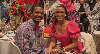A man and woman are sitting at a table, posing for a picture. The woman is wearing a pink dress and a hat. The man is wearing a plaid shirt and a bow tie. They are surrounded by other people sitting at tables. AI generated content