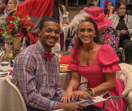A man and woman are sitting at a table, posing for a picture. The woman is wearing a pink dress and a hat. The man is wearing a plaid shirt. They are surrounded by other people sitting at tables. AI generated content