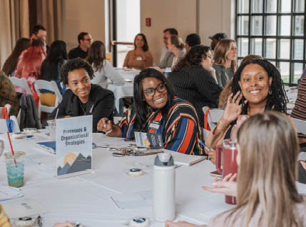 A group of people are sitting around a table, smiling and talking. There are several cups, a fork, and a knife on the table. A book is also visible on the table. The people are engaged in conversation and enjoying their time together. AI generated content
