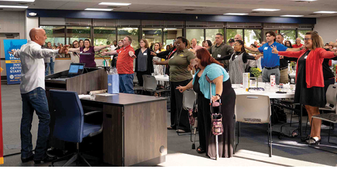 A group of people are standing in a room, some of them are clapping. A man is standing in front of a group of people, possibly giving a presentation or leading a meeting. There are chairs and a dining table in the room. AI generated content