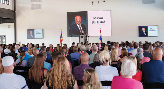 A large crowd of people is gathered in a room, watching a man speak at a podium. The man is wearing a suit and tie, and is standing in front of a large screen. The audience is seated in rows of chairs, and some of them are holding cell phones. The event appears to be a political rally, as the man speaking is a politician. AI generated content