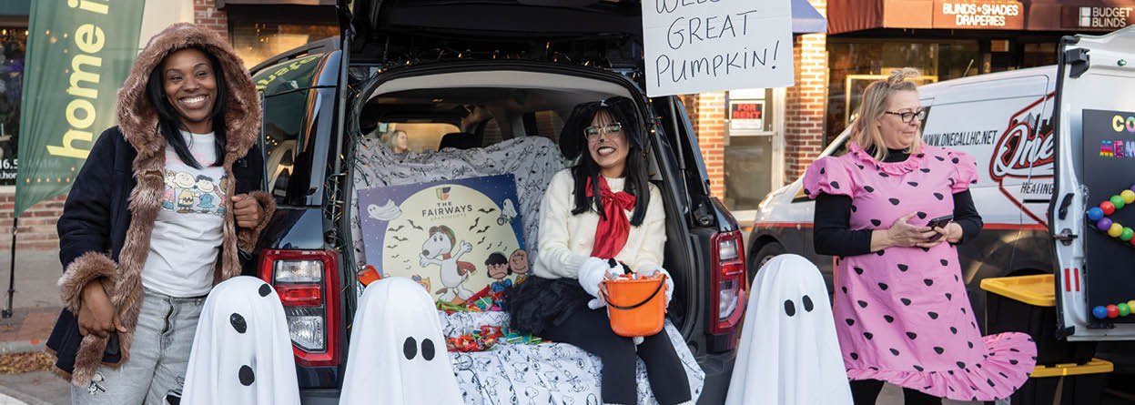 Three people are posing in front of a car with a sign that says “welcome great pumpkin". The car is decorated with Halloween decorations, including a jack-o-lantern and a ghost. The people are dressed in costumes, with one person wearing a ghost costume and the other two dressed in pumpkin costumes. AI generated content