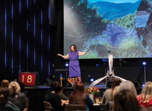 A woman in a purple dress is standing on a stage in front of a large screen. She is holding her arms out to the sides and appears to be performing for an audience. The audience is seated in chairs surrounding the stage, and there are several potted plants placed around the room. AI generated content