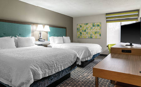A bedroom with a bed, desk, and TV. The bed has a blue headboard and a white comforter. The desk has a TV on it. AI generated content