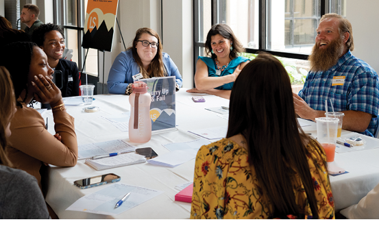 A group of people are sitting around a table, engaged in conversation. There are several cups, a bottle, and a book on the table. The people are of various ages and genders, and they appear to be enjoying their time together. AI generated content