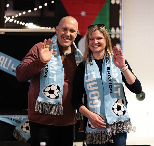 A man and woman are standing next to each other, both wearing blue scarves. The woman is wearing a scarf with a soccer ball on it, while the man is wearing a scarf with a soccer ball and the word “goal" on it. They are both waving and smiling. AI generated content