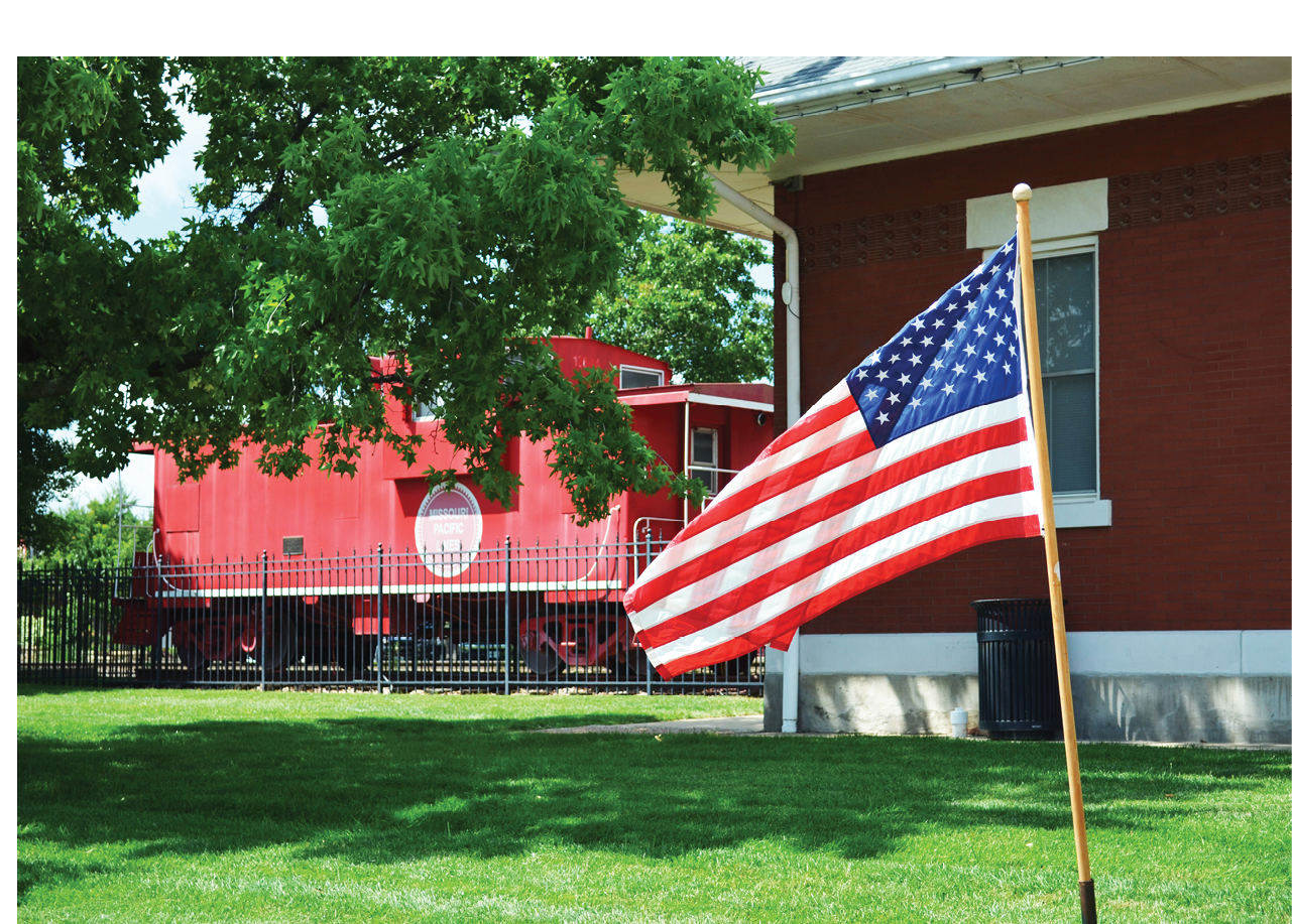 A red train is parked in front of a red brick building. An American flag is flying in front of the building. AI generated content