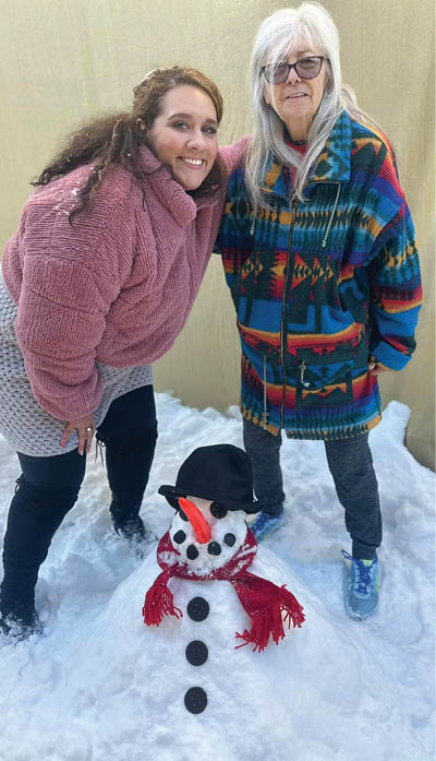 Two women pose for a picture in front of a snowman. AI generated content