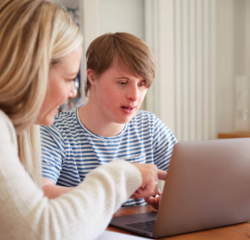 Downs Syndrome Man Sitting With Home Tutor Using Laptop For Lesson At Home