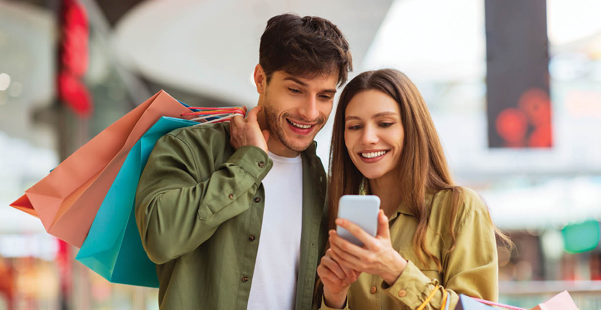Happy Couple Shopping Using Mobile Phone Application Holding Colorful Shopper Bags And Buying Clothes In Modern Mall On Weekend. Customers Hold Smartphone Browsing Internet For Sales. Ecommerce