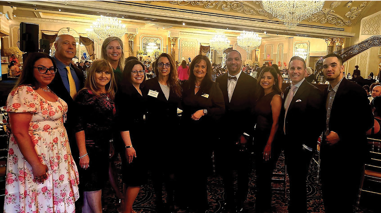 A group of people, both men and women, are standing together in a room. They are all dressed in formal attire, with some wearing ties. The group appears to be posing for a picture. AI generated content