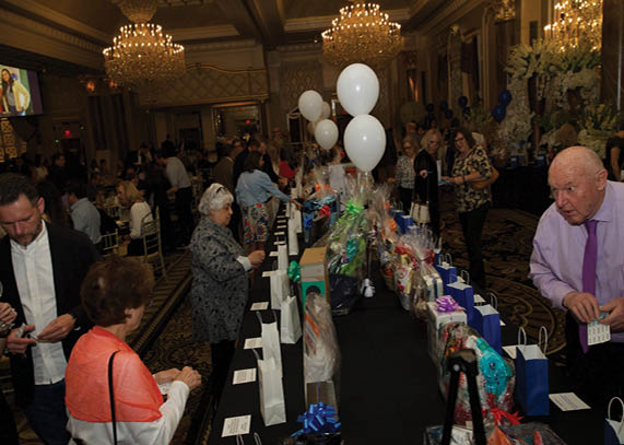 A large group of people are gathered in a room, socializing and enjoying a party. The room is filled with tables, some of which are covered with gift bags and balloons. The guests are standing around the tables, mingling and having a good time. AI generated content
