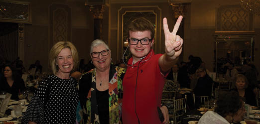 Three people pose for a photo in a banquet hall. A man in a red shirt is holding up a peace sign, while a woman in a polka dot shirt smiles. Another woman is also present in the scene. The banquet hall is filled with tables and chairs, and there are several wine glasses and cups placed on the tables. AI generated content