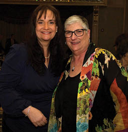 Two women are standing in a room, posing for a picture. One woman is wearing a black dress and the other is wearing a colorful jacket. There are several chairs in the room, some of which are placed around a dining table. There are also a few cups and a vase visible in the scene. AI generated content