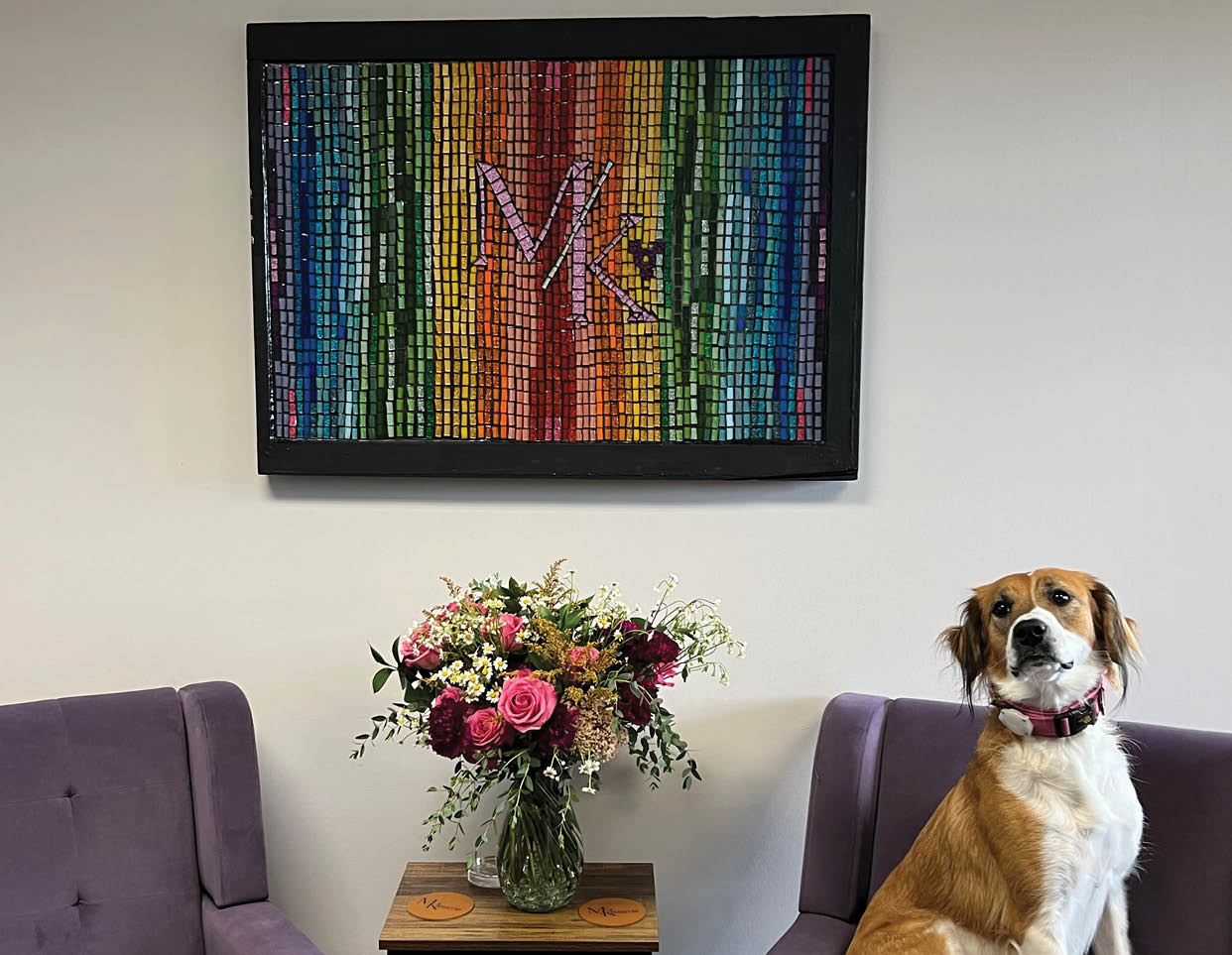 A dog sitting on a purple chair in front of a colorful picture. Description generated by AI