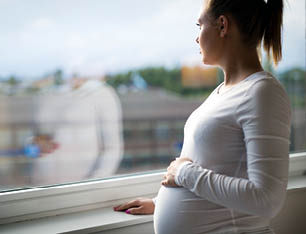 Thoughtful pregnant woman looks out the window home. Holding a hand on the tummy. Mental health and pregnancy.