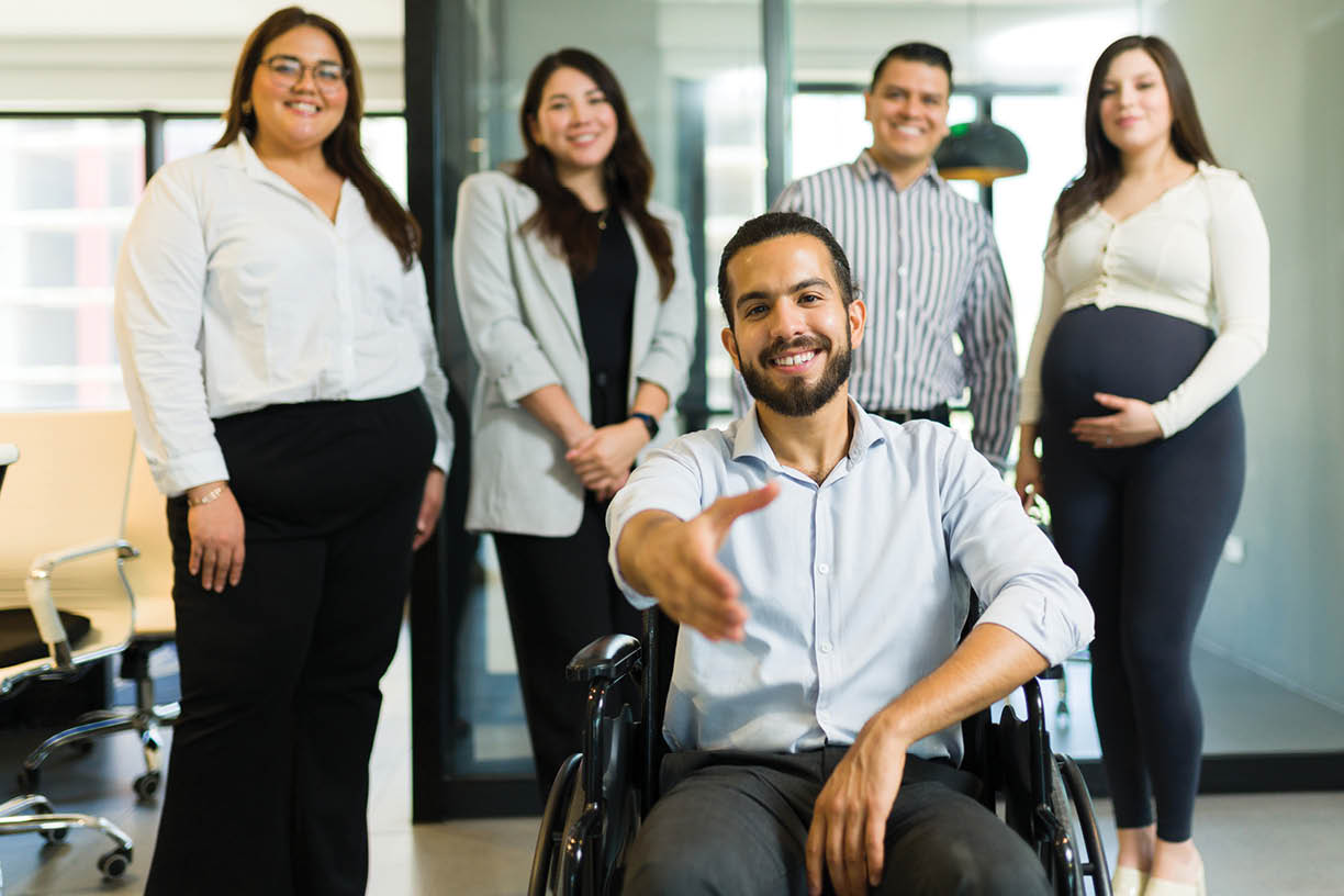 Portrait of a disabled manager sitting on wheelchair offering a handshake with his colleagues standing at back in office