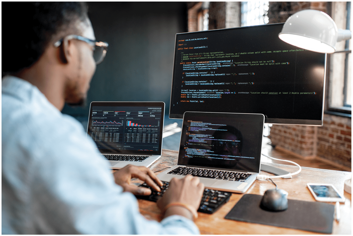 Young african male programmer writing program code sitting at the workplace with three monitors in the office. Image focused on the screen
