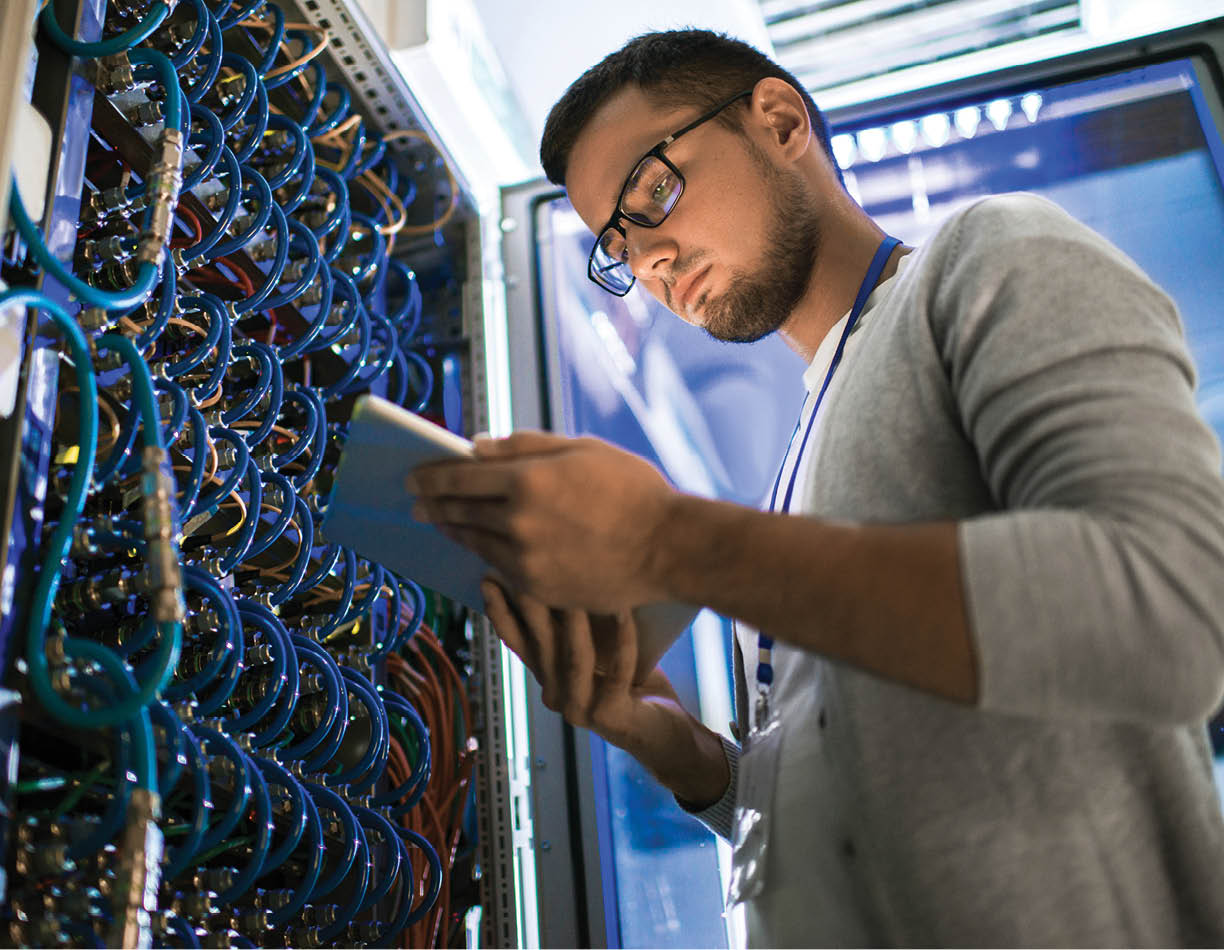 Low angle portrait of young man using digital tablet standing by server cabinet while working with supercomputer in blue light