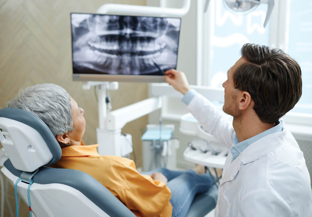 Back view of male dentist pointing at tooth X-ray image on screen during consultation in modern dental clinic