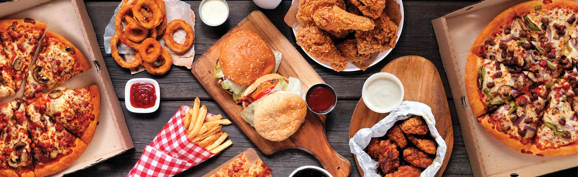 Table scene of assorted take out or delivery foods  Hamburgers, pizza, fried chicken and sides  Top down view on a dark wood banner background 